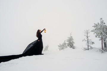 Young beautiful woman in long black dress with old lantern over winter hill background and snowfall. Fairy tale girl on polar winter landscape. Black witch in the snow.