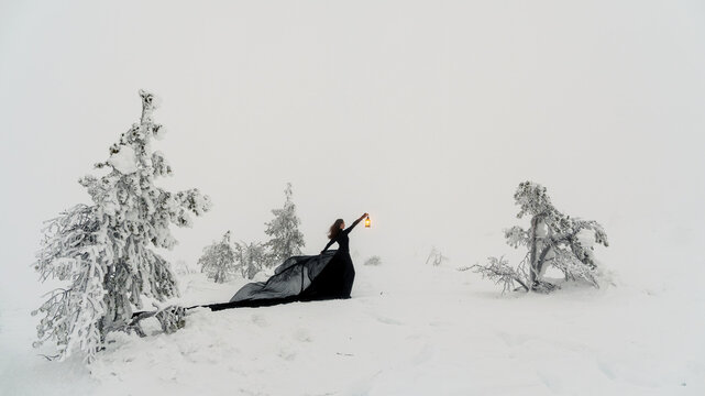 Young beautiful woman in long black dress with old lantern over winter hill background and snowfall. Fairy tale girl on polar winter landscape. Black witch in the snow. Panoramic view.