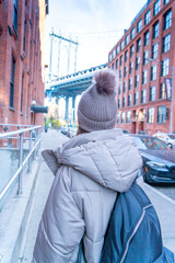 NEW YORK CITY WOMAN WEARING A WINTER HAT AND ENJOYING BROOKLYN BRIDGE  VIEW FROM DUMBO STREET. LIVING HAPPY DURING WINTER TIME IN UNITED STATES. TRAVEL, SUNNY DAY AND AMERICA CONCEPT.