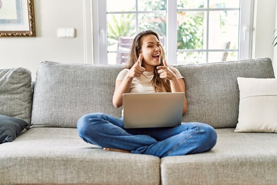 Beautiful young brunette woman sitting on the sofa using computer laptop at home pointing fingers to camera with happy and funny face. good energy and vibes.