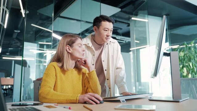 Crew multiracial team of creative people discussing a project on a computer looking at a monitor screen in modern office. Two young colleagues in casual asian man and woman point finger