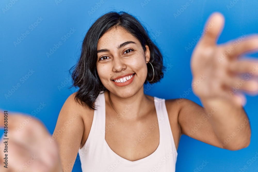 Wall mural Young hispanic woman standing over blue background looking at the camera smiling with open arms for hug. cheerful expression embracing happiness.