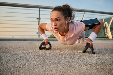 Determined female athlete sportswoman with earphones and smartphone holder exercising, doing press-ups push-ups, while practicing a bodyweight workout early in the morning on the city bridge