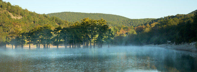 A delightful autumn sunrise over a mountain lake with cypresses growing out of the water. The surface of the water is covered with a milky mist dissolving in the air in the rays of the rising sun.