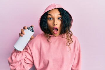 Young latin woman wearing sweatshirt holding graffiti spray scared and amazed with open mouth for...