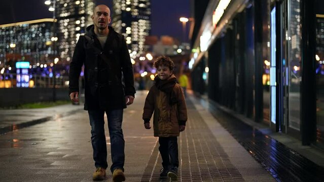 father and son walk along a blurry evening street against the background of city lights, then hold hands. the camera is moving. the general plan