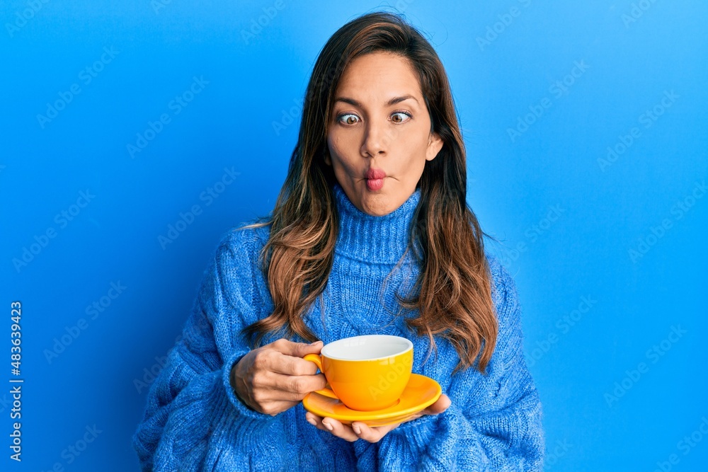 Sticker young latin woman drinking a cup of coffee making fish face with mouth and squinting eyes, crazy and