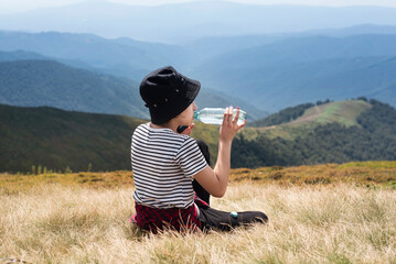 Young girl tourist sits on the grass in the mountains, rests after a long climb up and enjoys drinking water and talking on the phone with her parents about a successful vacation