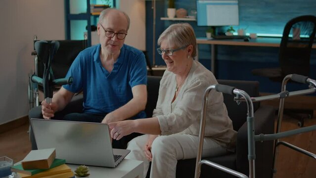 Couple with chronic disability giving high five after success on laptop, celebrating teamwork and retirement. Elder people with walking frame and crutches enjoying technology on computer.