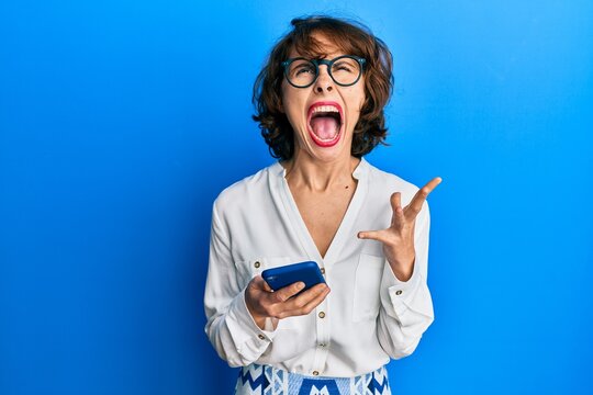 Young brunette woman using smartphone angry and mad screaming frustrated and furious, shouting with anger looking up.