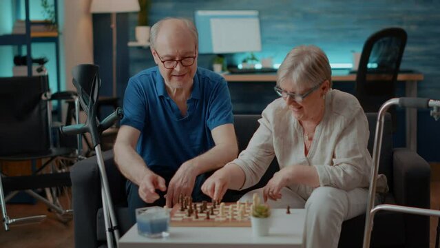 Elder couple with walk frame and crutches playing chess game on board. Retired people enjoying strategy competition with chess pieces to do checkmate and win intellectual match.