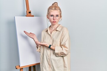 Beautiful caucasian woman with blond hair standing by painter easel stand presenting canva...