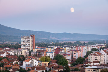 Fototapeta na wymiar Large moon over colorful Pirot cityscape during blue hour with purple clouds