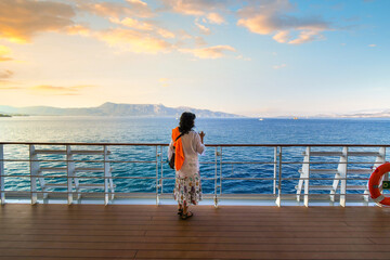 A woman stands on the deck of a cruise ship at sunset while it cruises the Mediterranean sea...