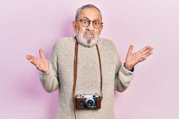 Handsome senior man with beard holding vintage camera clueless and confused with open arms, no idea...