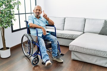 Fototapeta na wymiar Handsome senior man sitting on wheelchair at the living room showing and pointing up with finger number one while smiling confident and happy.