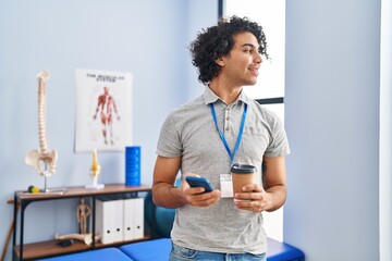 Young hispanic man physiotherapist using smartphone and drinking coffee at rehab clinic
