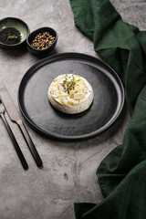 Oven baked camembert cheese with black pepper, sesame seeds on black plate, grey concrete surface. Homemade grilled brie with thyme, dark table setting