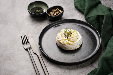 Oven baked camembert cheese with black pepper, sesame seeds on black plate, grey concrete surface. Homemade grilled brie with thyme, dark table setting