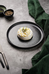 Oven baked camembert cheese with black pepper, sesame seeds on black plate, grey concrete surface. Homemade grilled brie with thyme, dark table setting