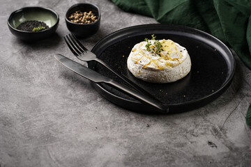 Oven baked camembert cheese with black pepper, sesame seeds on black plate, grey concrete surface. Homemade grilled brie with thyme, dark table setting