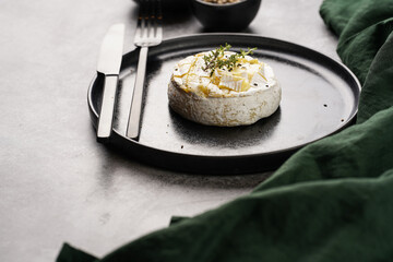Oven baked camembert cheese with black pepper, sesame seeds on black plate, grey concrete surface. Homemade grilled brie with thyme, dark table setting