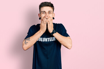 Young caucasian boy with ears dilation wearing volunteer t shirt laughing and embarrassed giggle covering mouth with hands, gossip and scandal concept