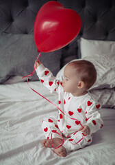 Beautiful little cute baby girl sitting on the bed and holding heart ballon. Valentine's Day.