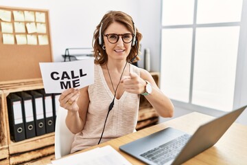 Middle age brunette woman wearing operator headset holding call me banner pointing fingers to...
