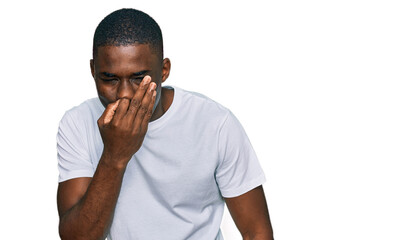 Young african american man wearing casual white t shirt smelling something stinky and disgusting, intolerable smell, holding breath with fingers on nose. bad smell