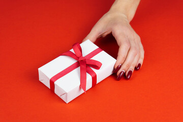 Woman hand hold white gift box with red bow on red table. Valentines day or birthday prsent concept