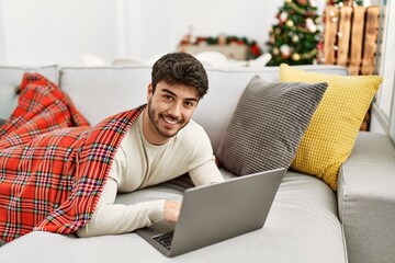 Young hispanic man smiling happy lying on the sofa using laptop at home.