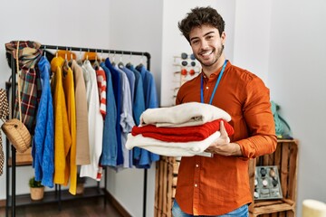 Young hispanic shopkeeper man smiling happy holding stack of sweater at clothing store.