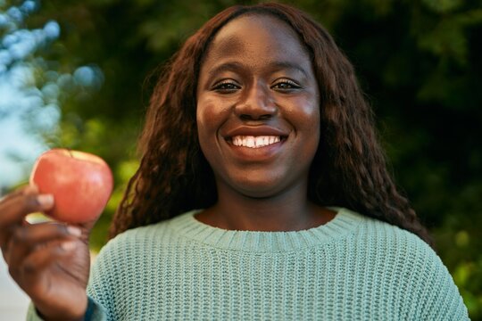 Young african woman smiling happy holding fresh red apple at the park