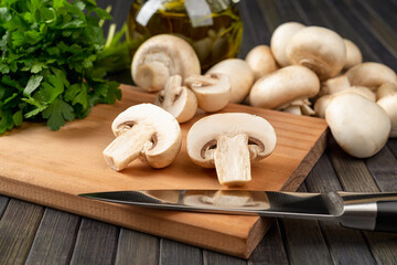 Raw champignon cut in half and and kitchen knife on a cutting board, whole button mushrooms and bunch of parsley over dark background. Vegetarian food ingredient, vegetable protein. - obrazy, fototapety, plakaty