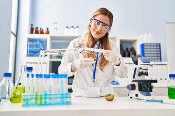 Young blonde woman wearing scientist uniform examining kiwi using magnifying glass at laboratory