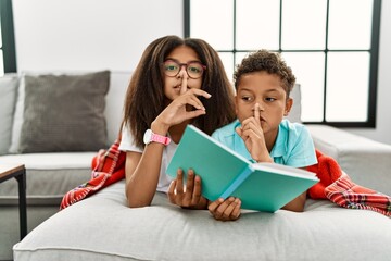 Two siblings lying on the sofa reading a book asking to be quiet with finger on lips. silence and secret concept.