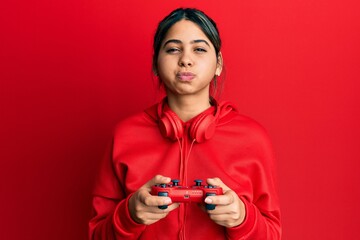 Young latin woman playing video game holding controller puffing cheeks with funny face. mouth...