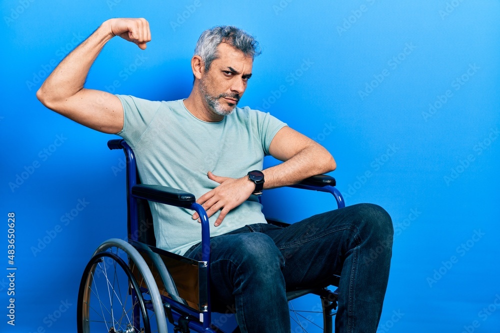 Poster handsome middle age man with grey hair sitting on wheelchair strong person showing arm muscle, confi