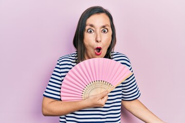 Obraz na płótnie Canvas Middle age hispanic woman waving hand fan cooling air in summer scared and amazed with open mouth for surprise, disbelief face