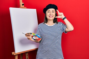 Middle age hispanic woman standing drawing with palette by painter easel stand smiling and confident gesturing with hand doing small size sign with fingers looking and the camera. measure concept.