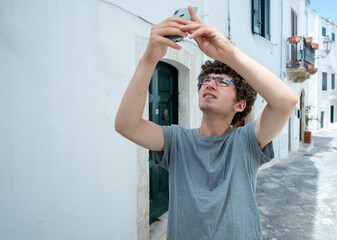 Ostuni, Puglia, Italy. August 2021. A young Caucasian is busy taking pictures of him with his mobile phone in one of the white alleys of the historic center of the village. Blurred background.