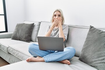 Young caucasian woman using laptop at home sitting on the sofa shocked covering mouth with hands...