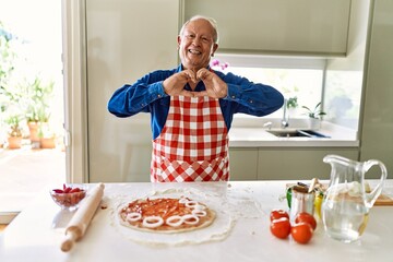 Senior man with grey hair cooking pizza at home kitchen smiling in love showing heart symbol and shape with hands. romantic concept.