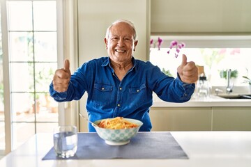 Senior man with grey hair eating pasta spaghetti at home approving doing positive gesture with hand, thumbs up smiling and happy for success. winner gesture.