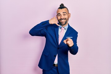 Young hispanic man wearing business suit and tie smiling doing talking on the telephone gesture and pointing to you. call me.