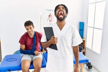 Young hispanic man working at pain recovery clinic with a man with broken arm angry and mad screaming frustrated and furious, shouting with anger. rage and aggressive concept.