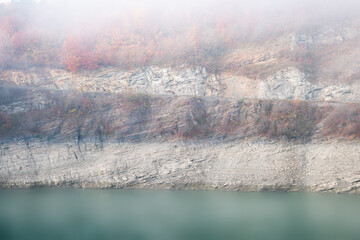 Scenic, glowing, colorful view of long exposure lake surface, rocky cliff, mountain road and autumn colored trees on a misty, moody day