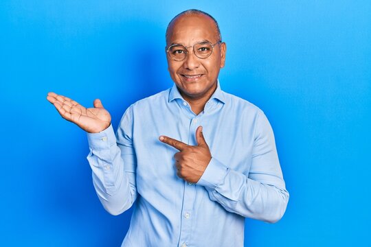Middle age latin man wearing casual clothes and glasses amazed and smiling to the camera while presenting with hand and pointing with finger.