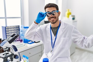 Young hispanic man working at scientist laboratory wearing magnifying glasses celebrating achievement with happy smile and winner expression with raised hand